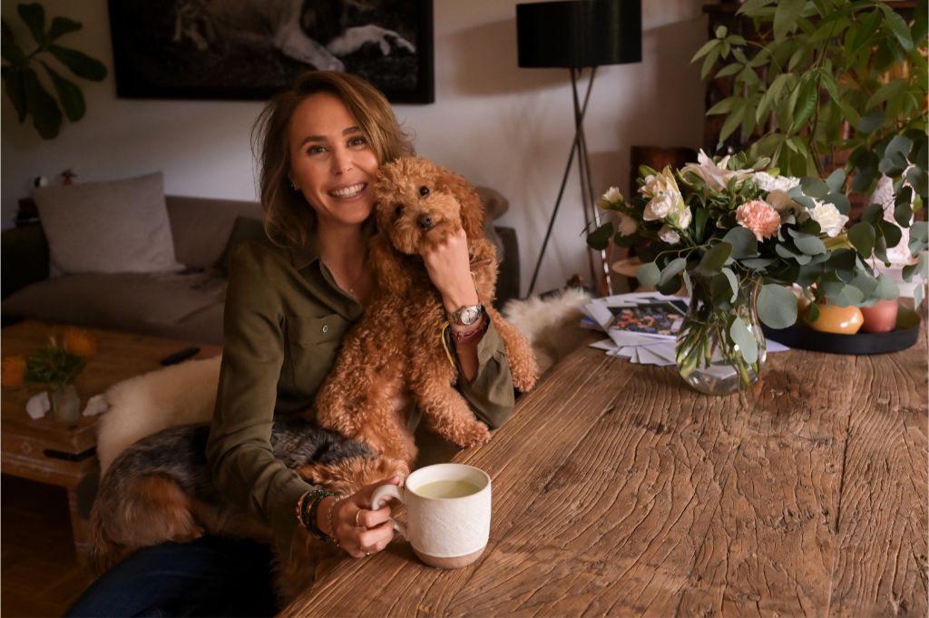 Founder sitting at a wooden table holding a dog and a cup of tea at home

